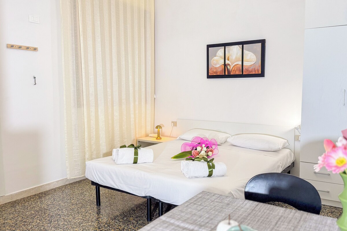 Ein Hotelzimmer mit einem Bett, einem Nachttisch mit Lampe, einem gerahmten Bild an der Wand, einem Kleiderschrank und einer Vase mit Blumen.