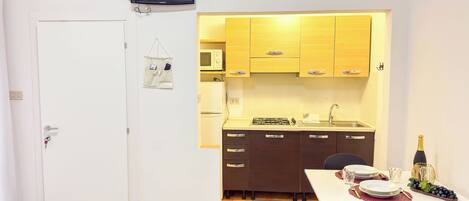 Una cocina compacta con armarios de madera, microondas y cocina a gas. Hay un comedor con una mesa lista para dos personas, una botella de champán y una bandeja de fruta.