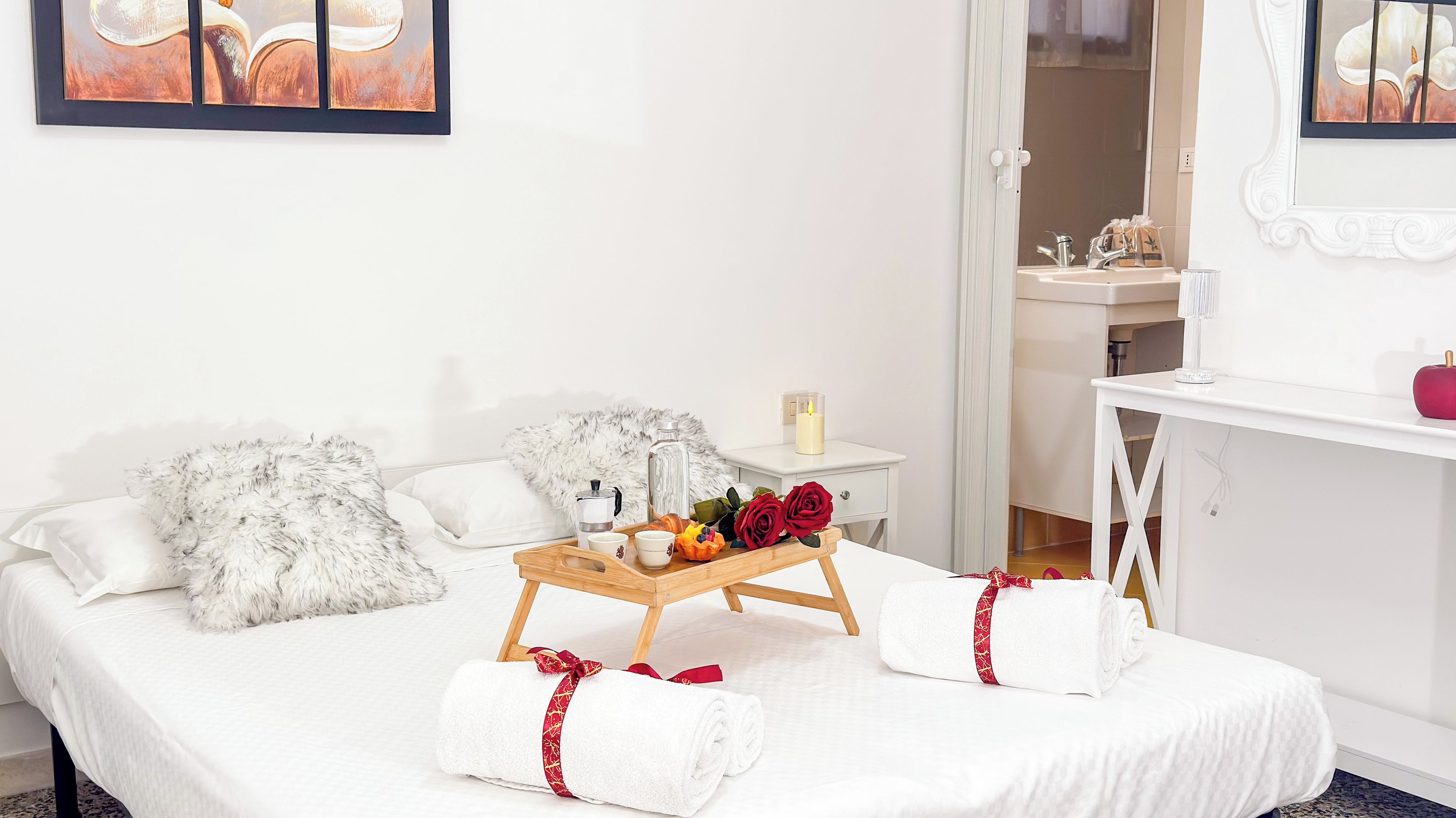 Un lit bien fait, agrémenté de coussins moelleux, une table de chevet avec une bougie, un plateau avec des fleurs et un service à thé, et un miroir au-dessus du lavabo.