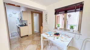 A compact kitchen with a stove, sink, and a small refrigerator. A dining table with chairs is set in a bright room with a window and potted plants. - Cà 4 nevodi Chiara by Ca-Sa Check (Chioggia)