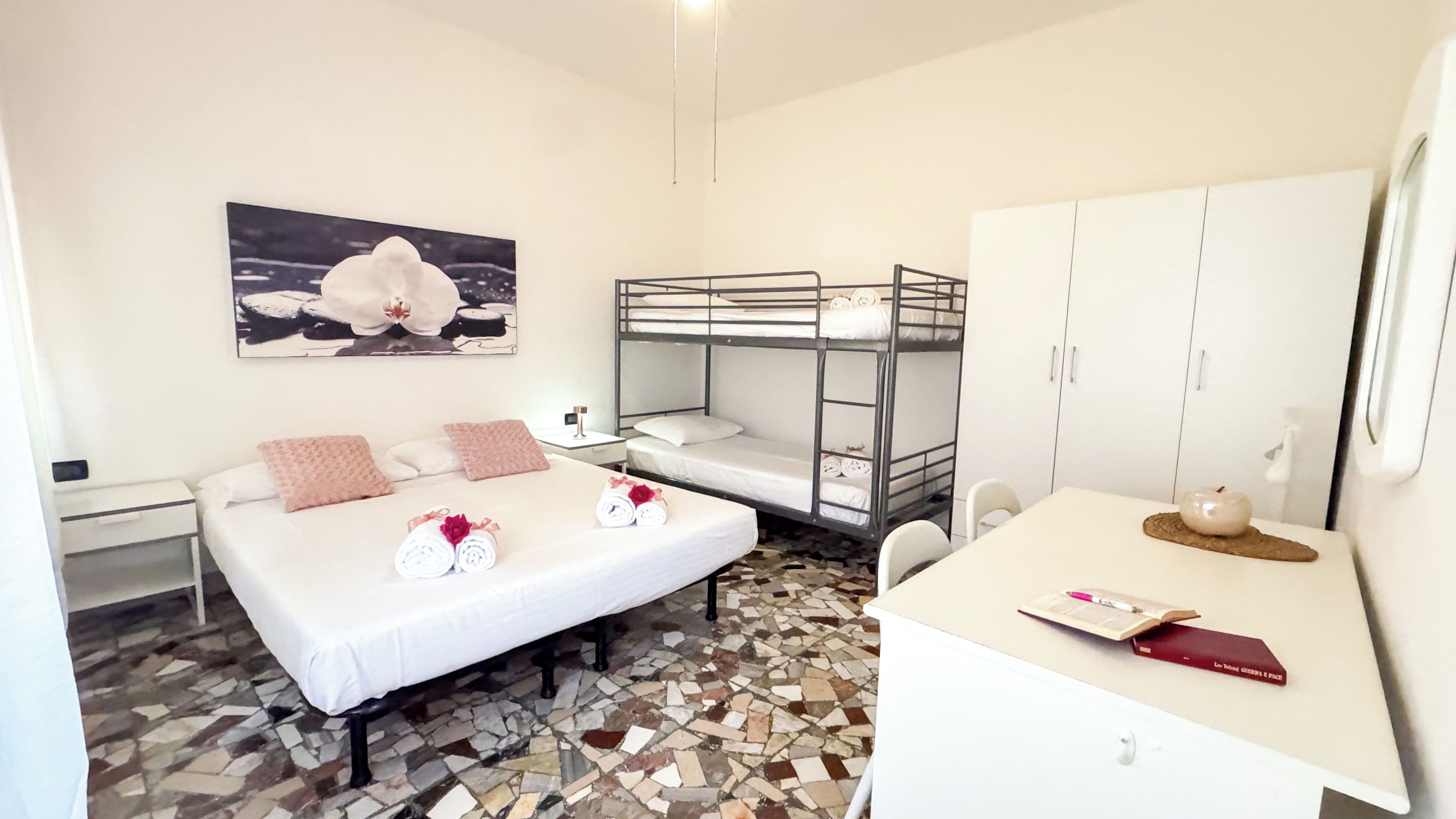 Habitación de hotel con cama, literas, escritorio, silla, pequeña mesa, lámpara, un cuadro y suelo de baldosas.