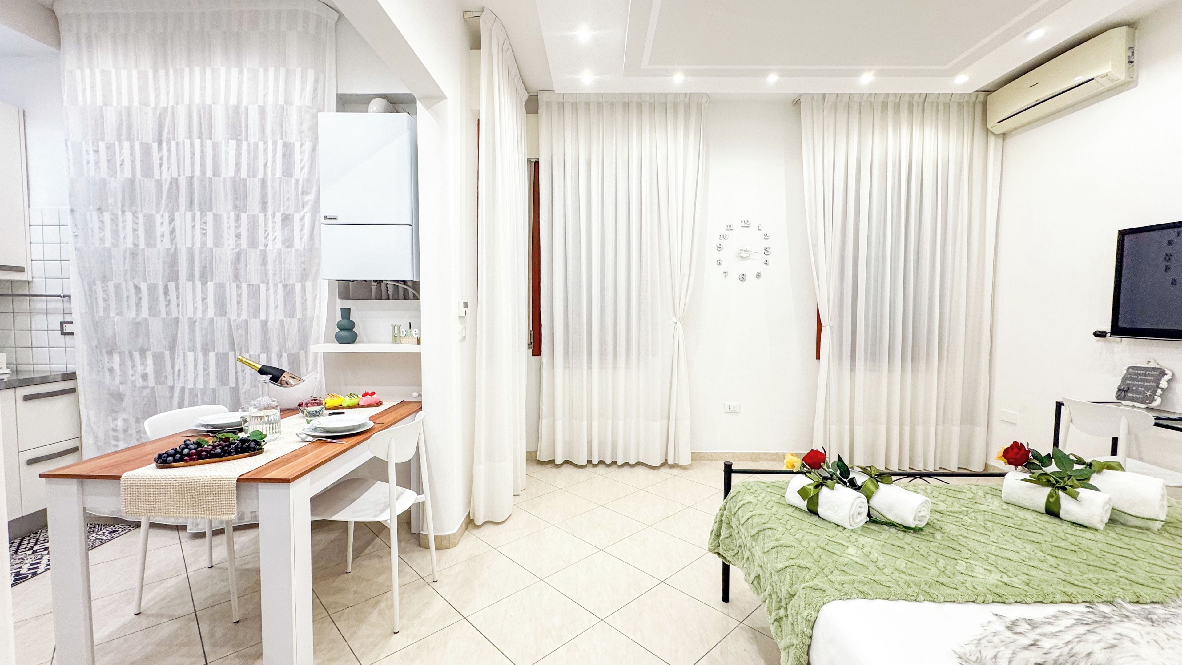Habitación de hotel moderna con zona de comedor, una cama con manta verde y un televisor montado en la pared.