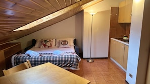 A small, cozy room with a sloped ceiling, a bed with plaid bedding, a wooden table, and a kitchenette area. - Baradello Apartment & Flat by ca-Sa Check  (Corteno Golgi)
