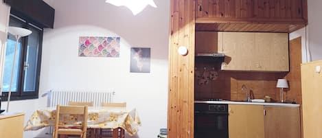 A compact kitchen with wooden cabinets, a built-in oven, and a sink. There is a dining table with chairs and a window with blinds.
