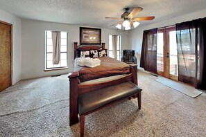 A bedroom with a bed, a bench, a ceiling fan, a massage chair, and a large window with curtains. - Cozy Cove Lodge | Game Room + Private Hot Tub! (Ruidoso)