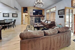 A spacious living room with a stone fireplace, a large sofa, a dining area with a wooden table and chairs, and a television mounted on the wall. - Cozy Cove Lodge | Game Room + Private Hot Tub! (Ruidoso)