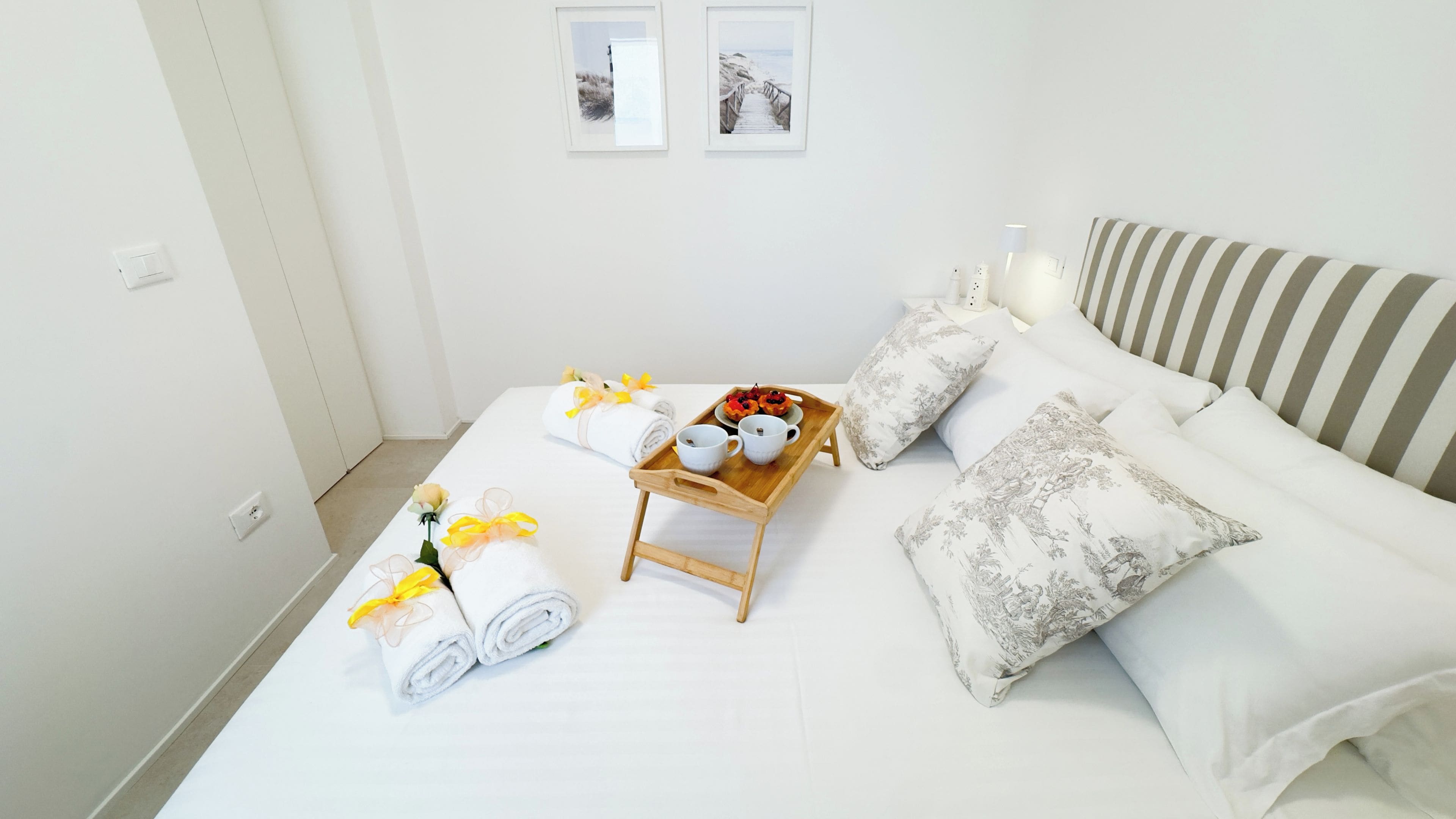A neatly made bed with white linens, a wooden tray with cups and a plate of food, and two rolled towels with yellow ribbons.