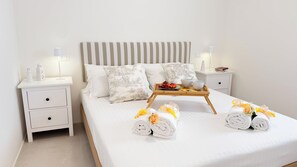 Ein ordentlich bezogenes Bett mit weißen Laken, ein Tablett mit Früchten und Schalen sowie aufgerollte Handtücher mit Blumen.
