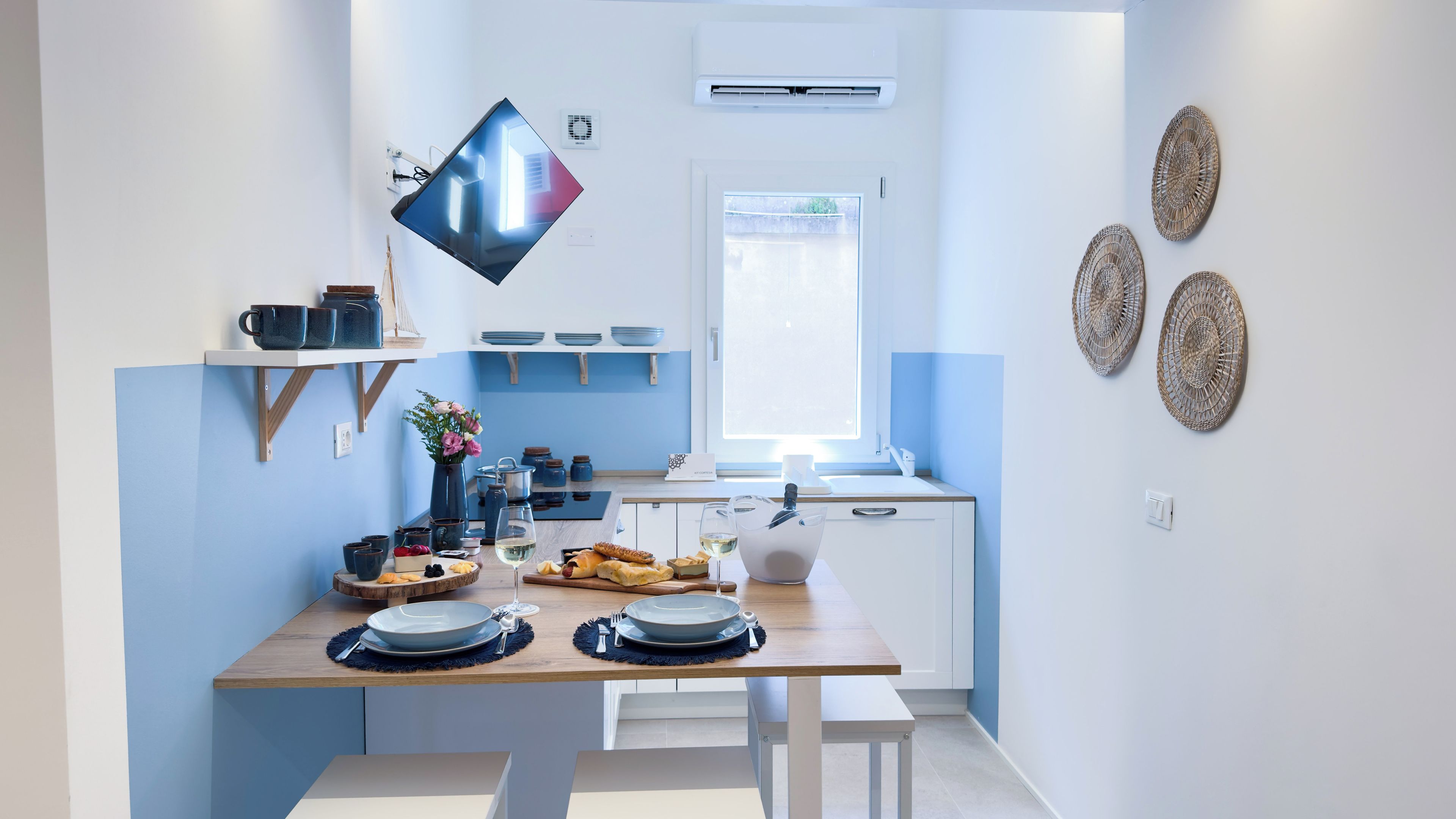 Une cuisine moderne avec une table à manger pour deux, un téléviseur fixé au mur et des œuvres d’art décoratives en fibres tressées.