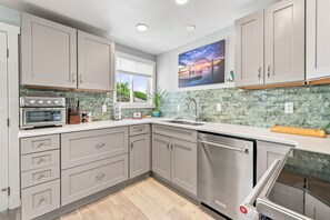 A modern kitchen with grey cabinets, a stainless steel dishwasher, and a window with a view of a bridge. - Coastal Cottage Near Yaquina Views, Sleeps 6 (Newport)