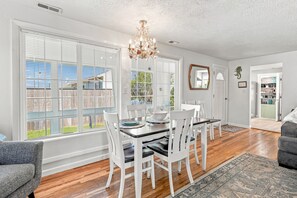 A dining area with a table set for four, white chairs, a chandelier, and large windows with blinds. - Coastal Cottage Near Yaquina Views, Sleeps 6 (Newport)