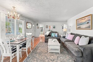Ein Wohnzimmer mit grauem Sofa, einem Couchtisch, einer Chandelierleuchtre und einem Essbereich mit Tisch und Stühlen.