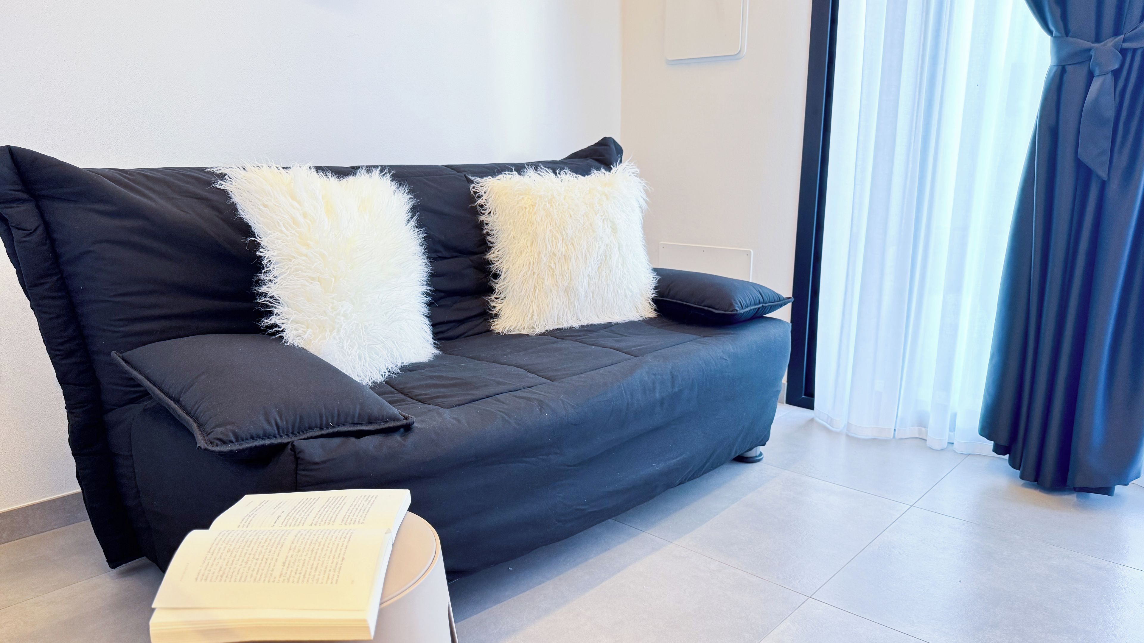 Una sala de estar moderna con un sofá negro, dos almohadas blancas mullidas y un libro sobre una mesita.