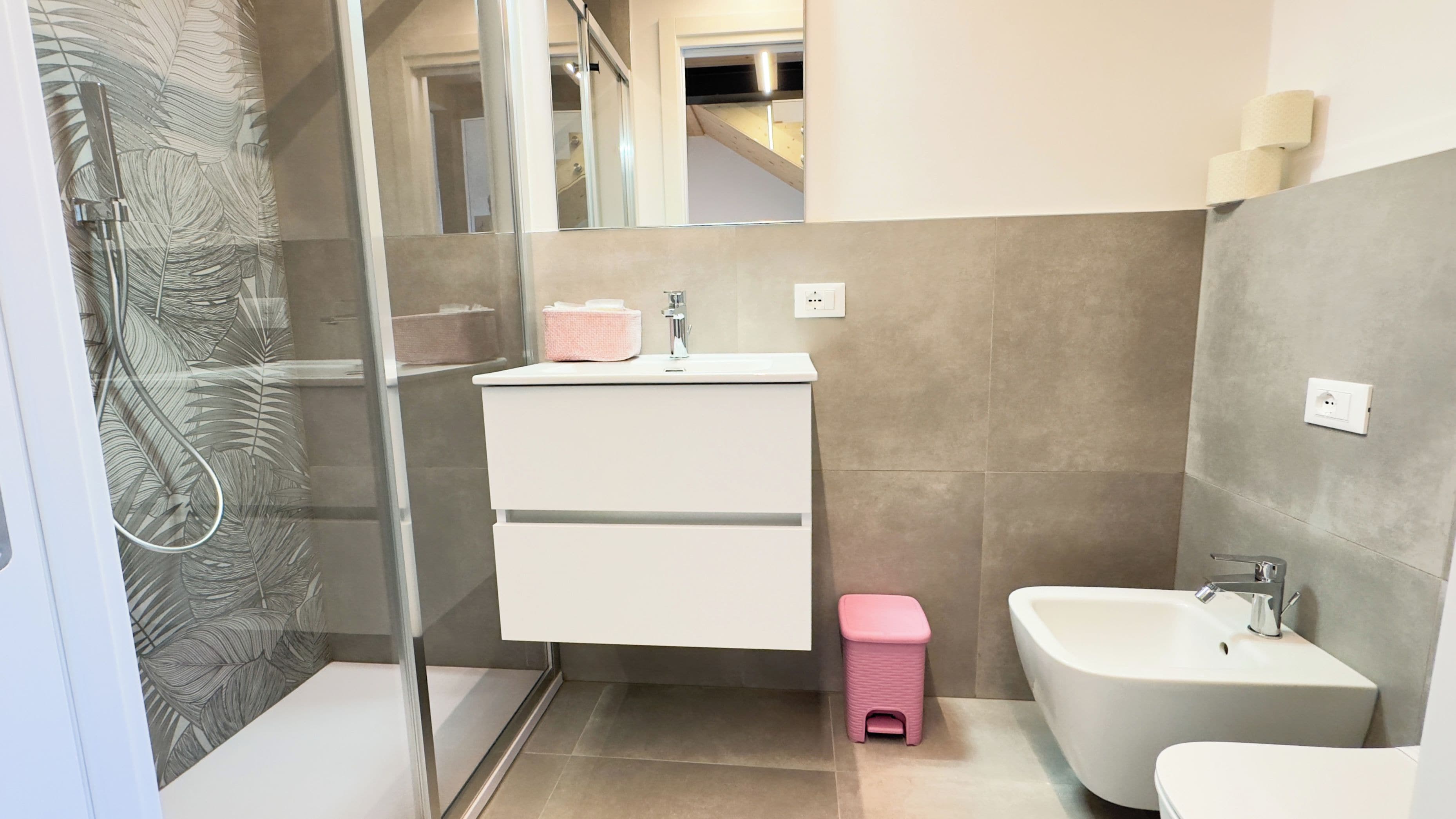 Um banheiro moderno com box de vidro, pia branca e um dosador de sabonete rosa, e também um bidê branco.