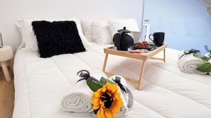 Uma cama bem arrumada com roupa de cama branca, um travesseiro preto, uma pequena mesa de cabeceira de madeira com um bule, uma xícara e um tabuleiro com comida.