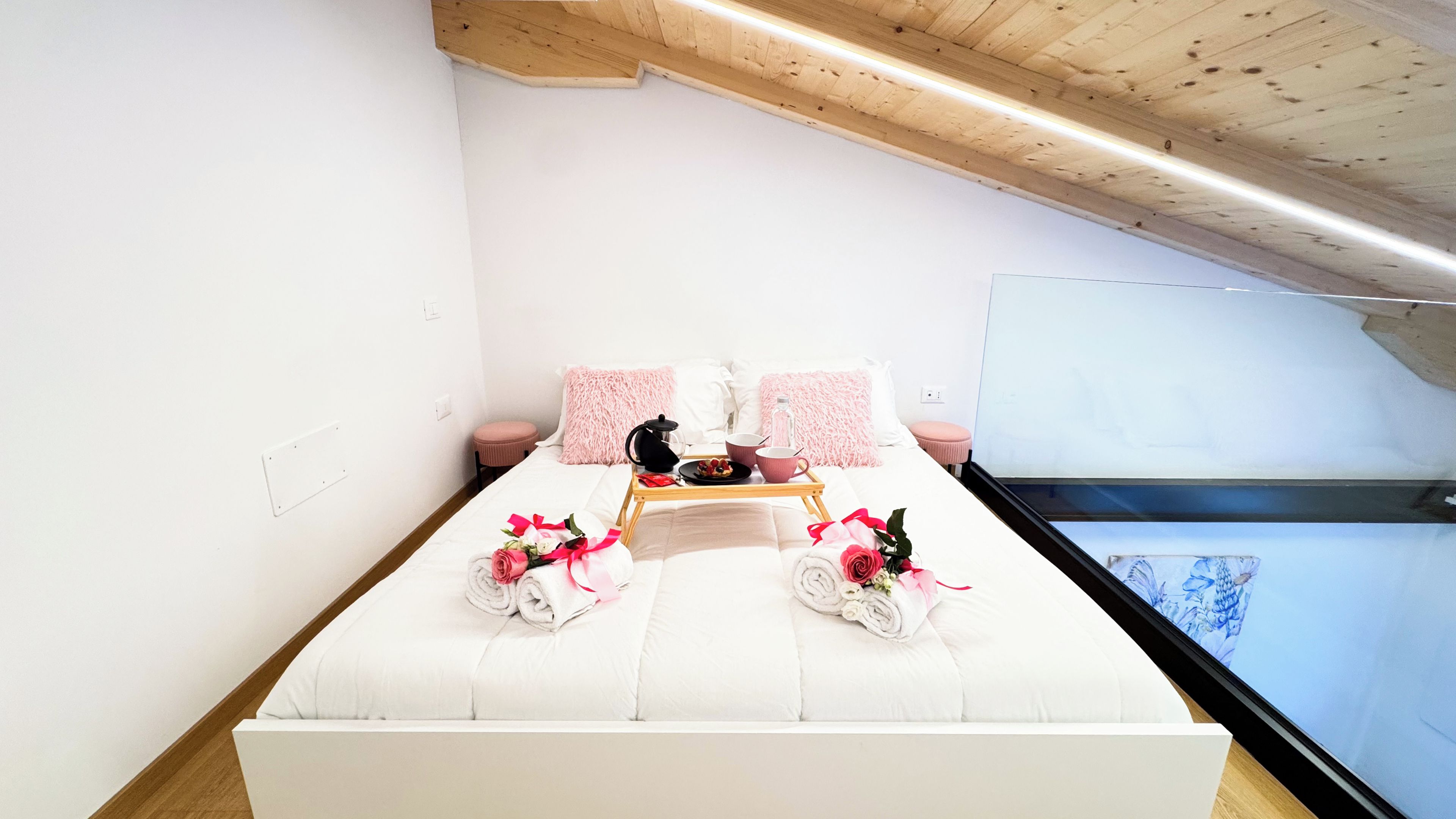 Uma cama bem arrumada com roupa de cama rosa e branca, um tabuleiro com itens de pequeno-almoço, e uma janela com vista para um céu azul e nuvens.