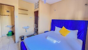 Ein Hotelzimmer mit einem Bett, einem Deckenventilator, einem Badezimmer mit Waschbecken und Toilette sowie einem kleinen Regal.