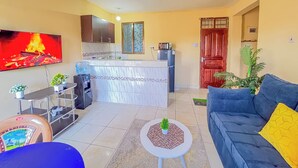 Ein Wohnzimmer mit blauem Sofa, Fernseher, Küchenzeile inklusive Kühlschrank und Deckenventilator.