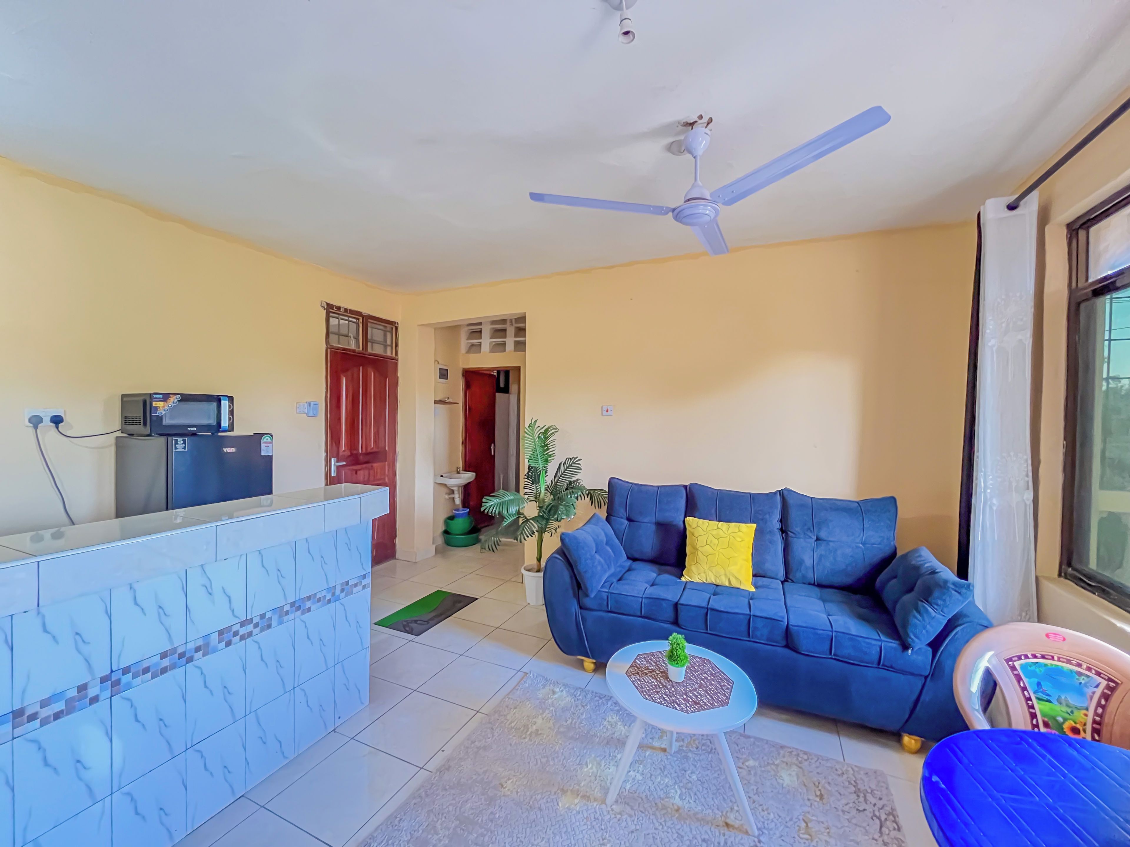 Una sala de estar con un sofá azul, una mesita redonda, un ventilador de techo y una zona de cocina con un microondas en un mueble.