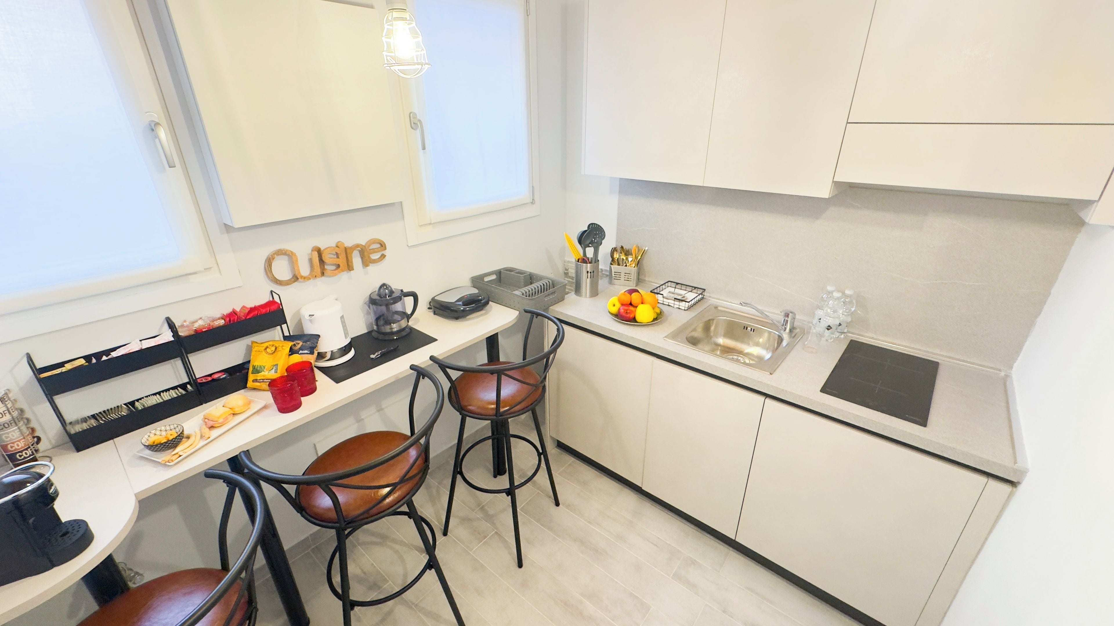 Una kitchenette compacta con cafetera, hervidor y frutero. Hay dos taburetes y una mesa pequeña.