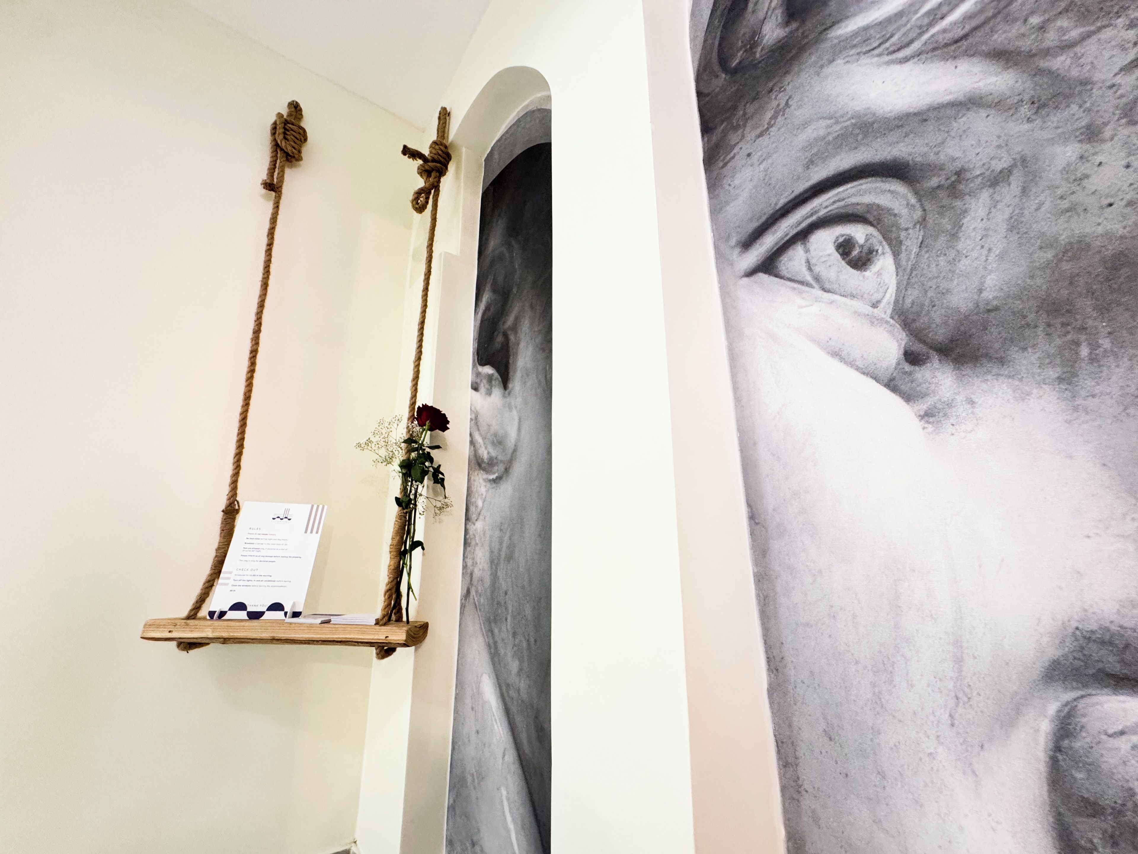 Quarto com prateleira de madeira suspensa por cordas, quadro emoldurado e parte de uma parede com um grande mural detalhado.