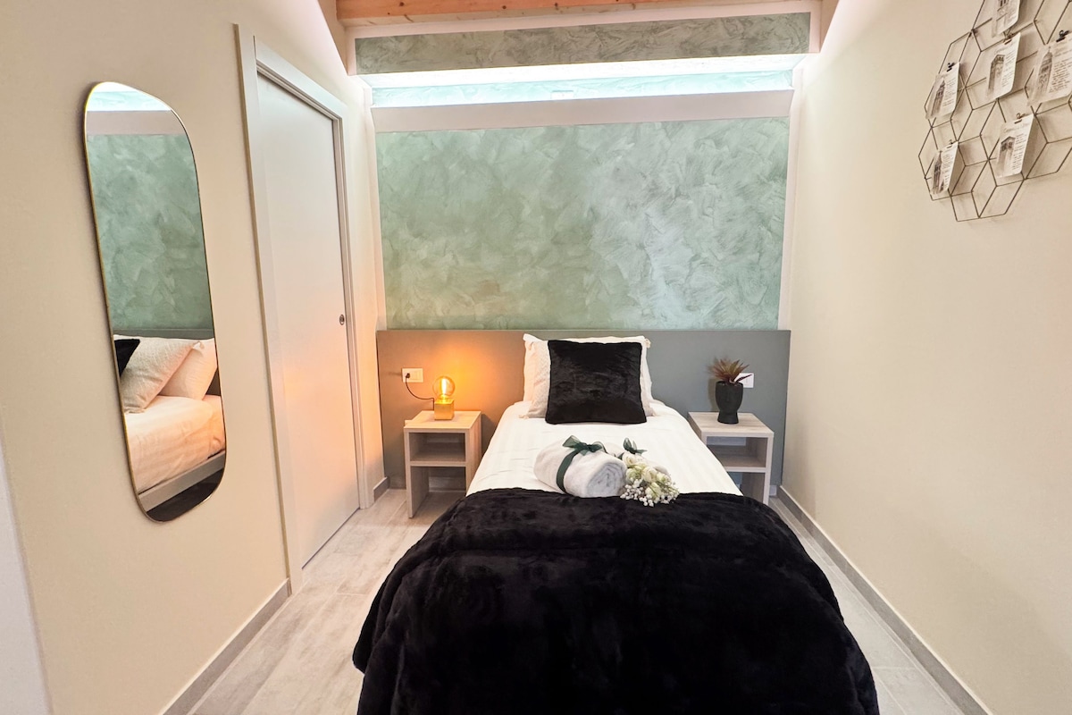 Ein schmaler Flur, der zu einem Schlafzimmer mit Bett, Nachttisch, Lampe und einer Wanddekoration führt.