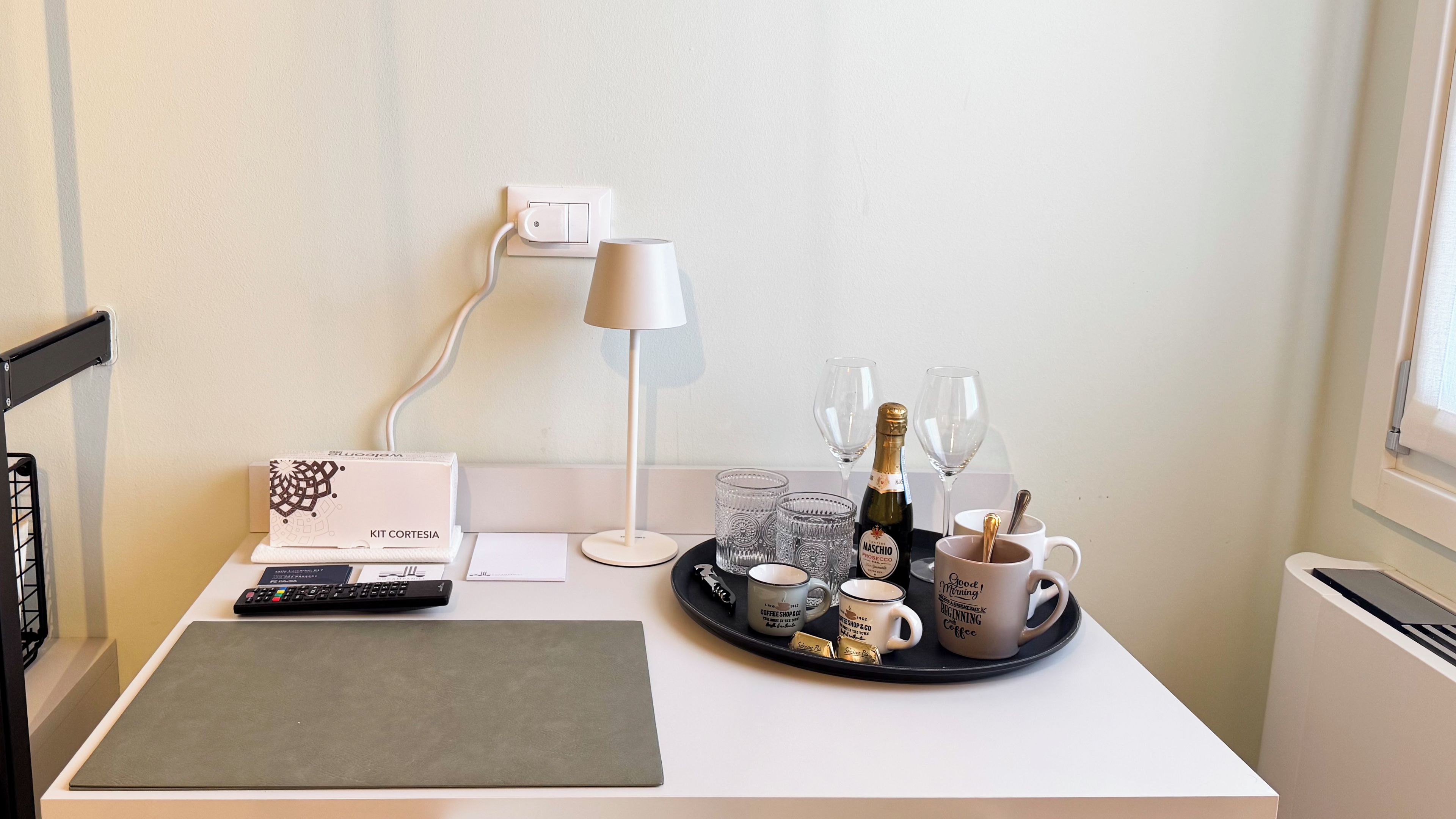 A neatly arranged workspace with a desk, a keyboard, a lamp, a tray with coffee mugs, glasses, and a bottle of champagne.