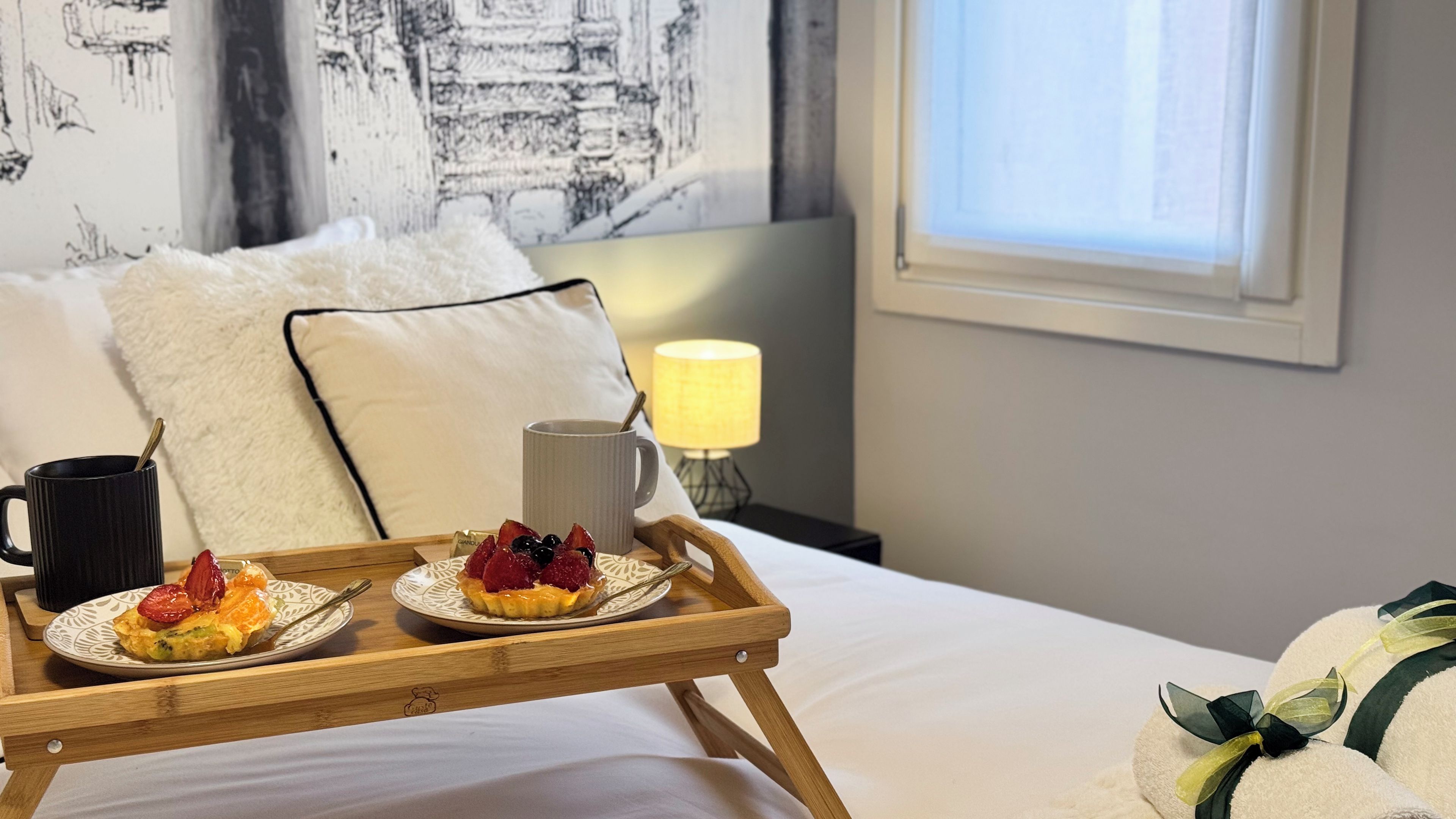 Una bandeja de desayuno bien dispuesta con un croissant, fruta y una taza sobre una cama con ropa blanca, almohadas y una lámpara en una habitación con ventana y una obra de arte en la pared.
