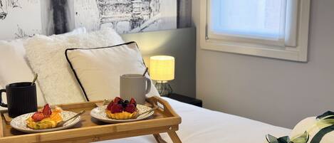Una bandeja de desayuno bien dispuesta con un croissant, fruta y una taza sobre una cama con ropa blanca, almohadas y una lámpara en una habitación con ventana y una obra de arte en la pared.