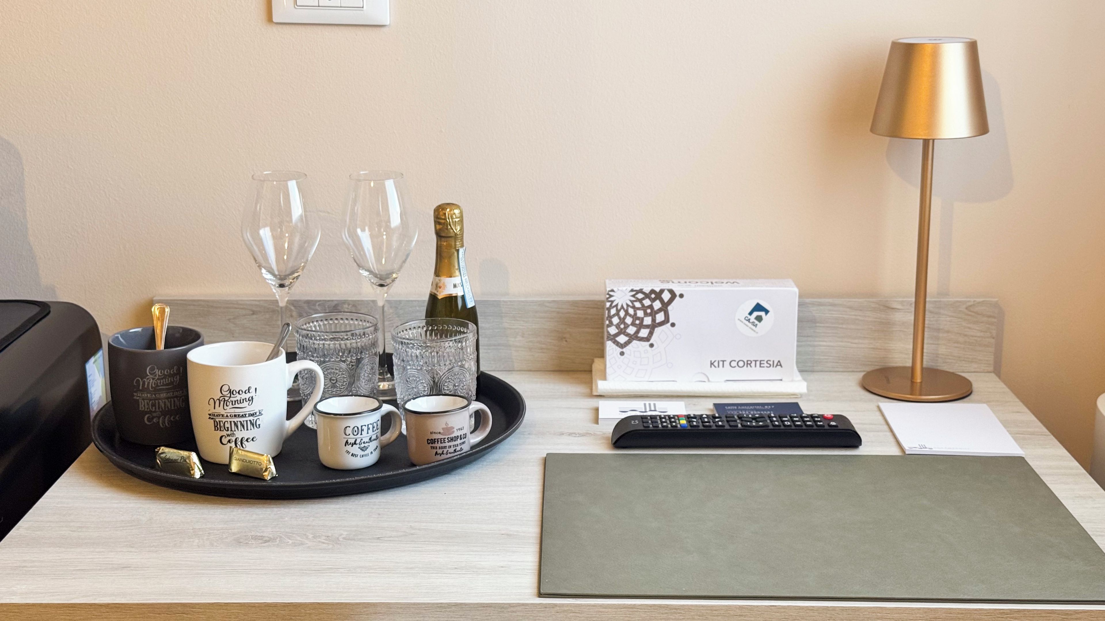 Ein Hotelzimmer mit einem Tablett mit Kaffeetassen, Gläsern, einer Champagnerflasche, einer Fernbedienung, einer mit 'KIT CORTESIA' beschrifteten Box und einer Lampe.