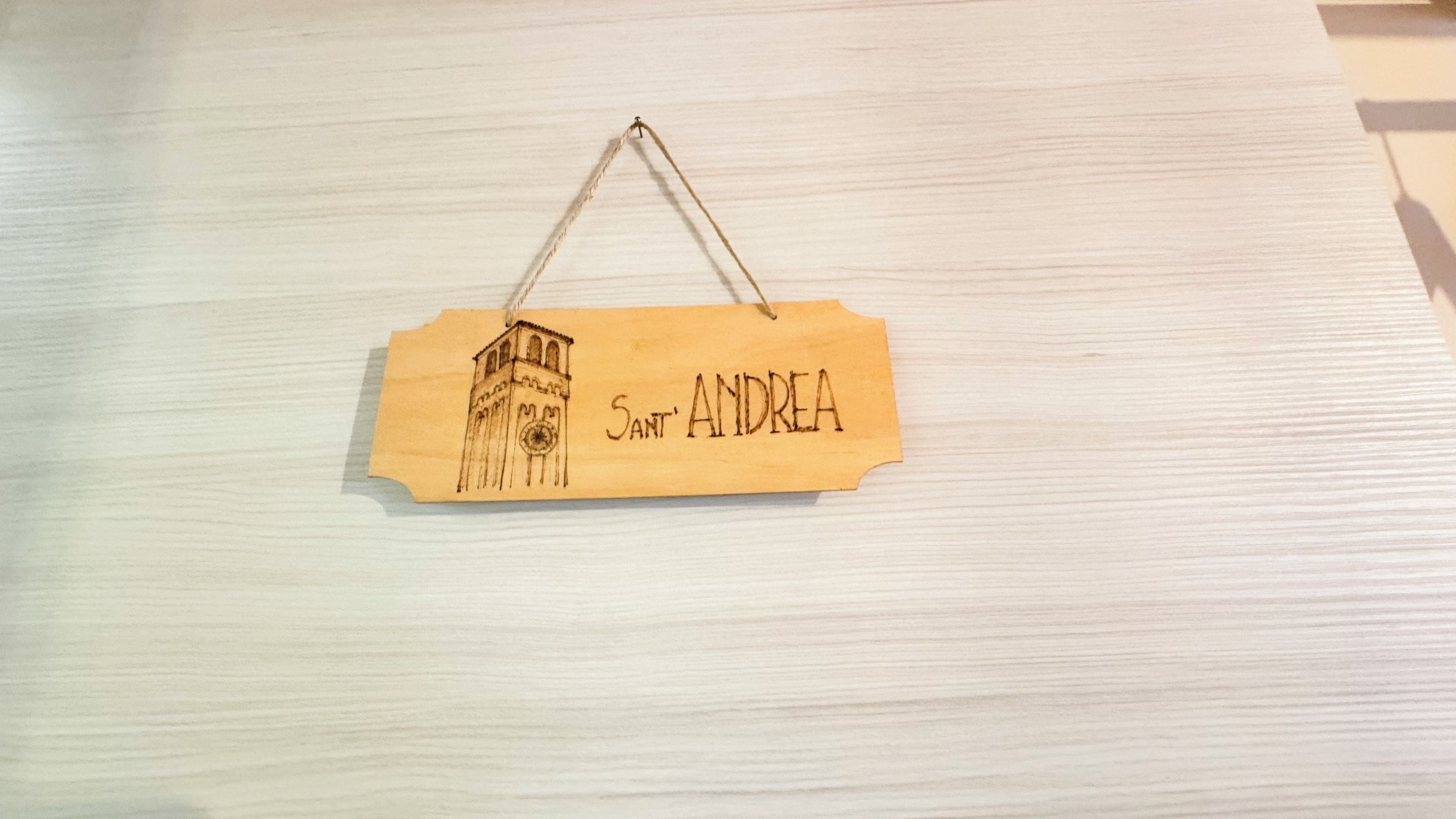 A wooden sign with a sketch of a tower and the name 'Sant' Andrea' on a light wooden surface.