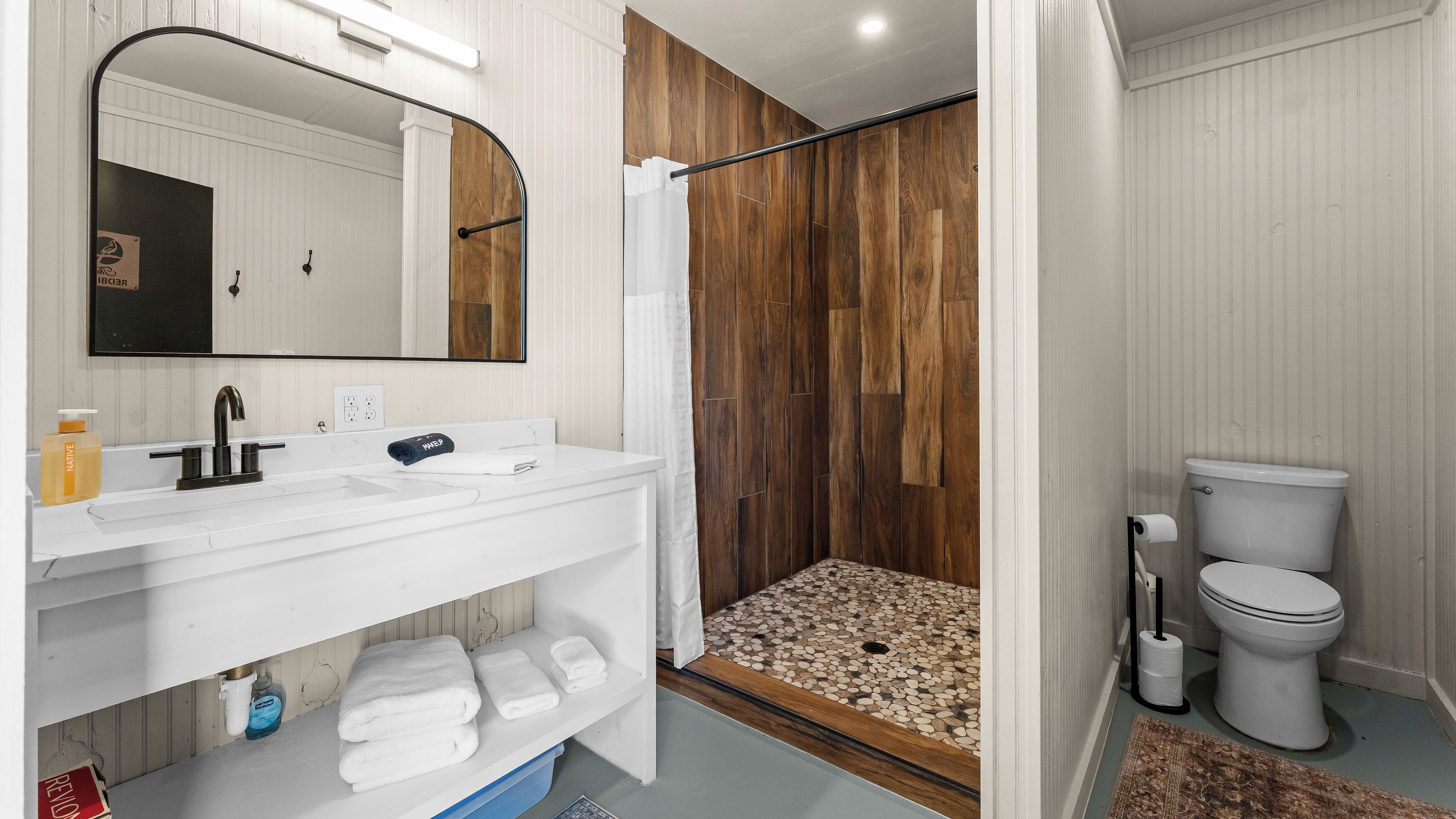 A modern bathroom with a white vanity, a large mirror, a shower with a wooden panel wall, and a toilet.