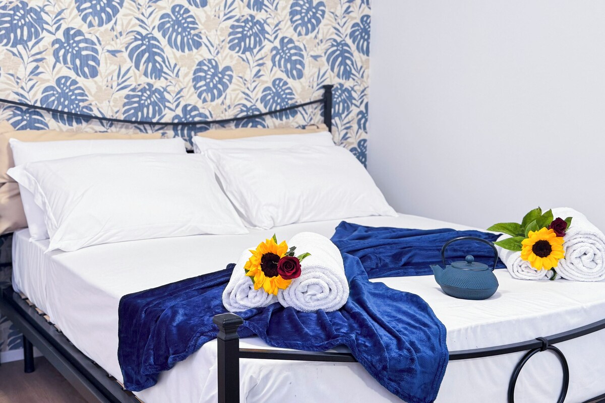 A neatly made bed with white linens, a blue throw blanket, and two towels with sunflowers and roses.