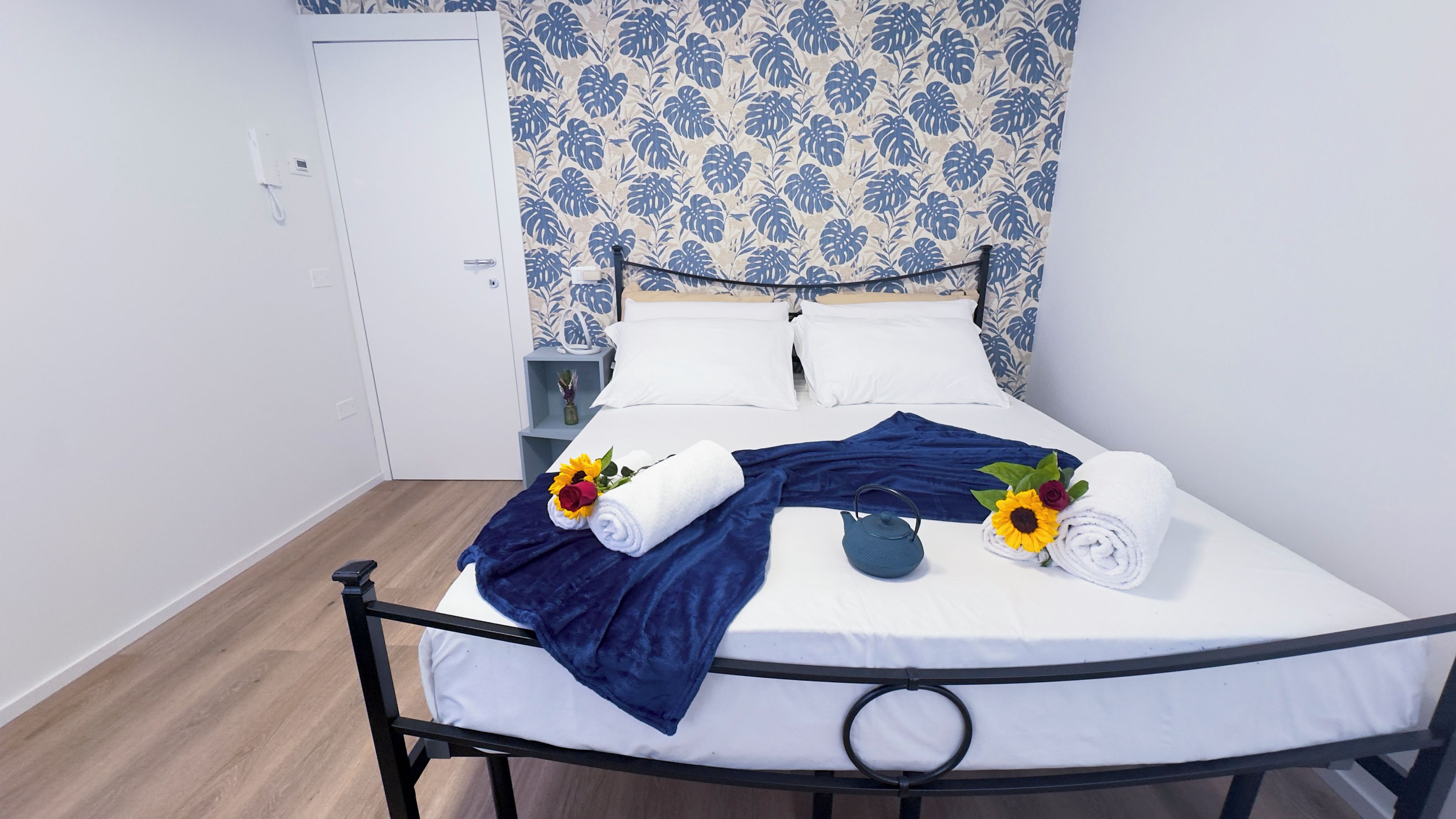 Un letto rifatto con lenzuola bianche, una coperta azzurra e asciugamani decorati con girasoli e rose.
