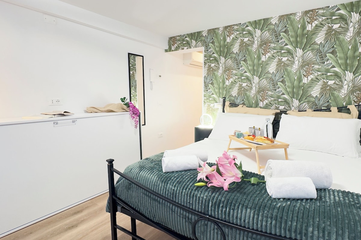 A modern bedroom with a bed, a bedside table with a tray of breakfast, a mirror, and a tropical leaf-patterned wall.