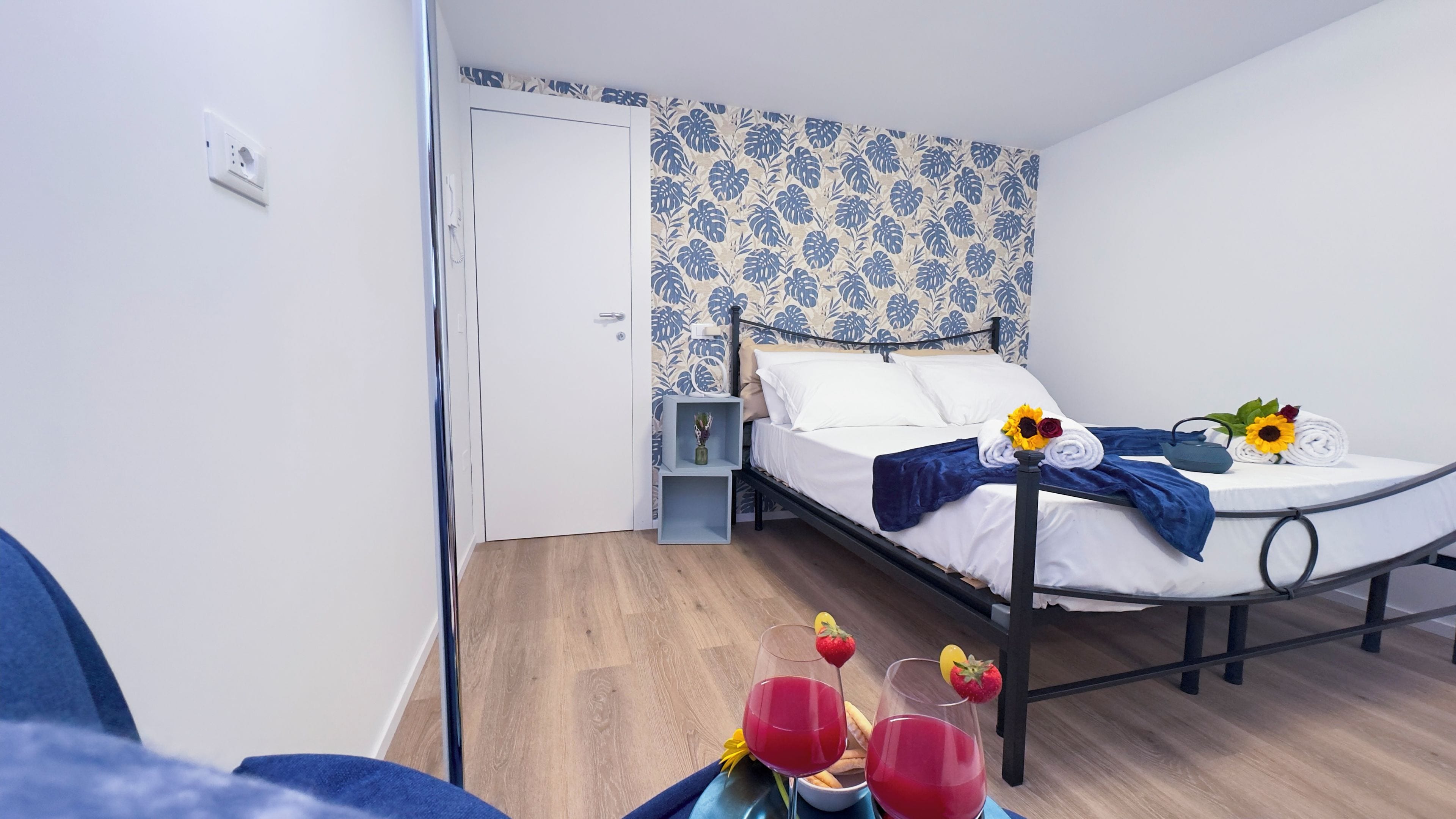 Una cama cuidadosamente hecha con una manta azul, una mesita de noche con una planta y una pared con estampado floral azul y blanco.