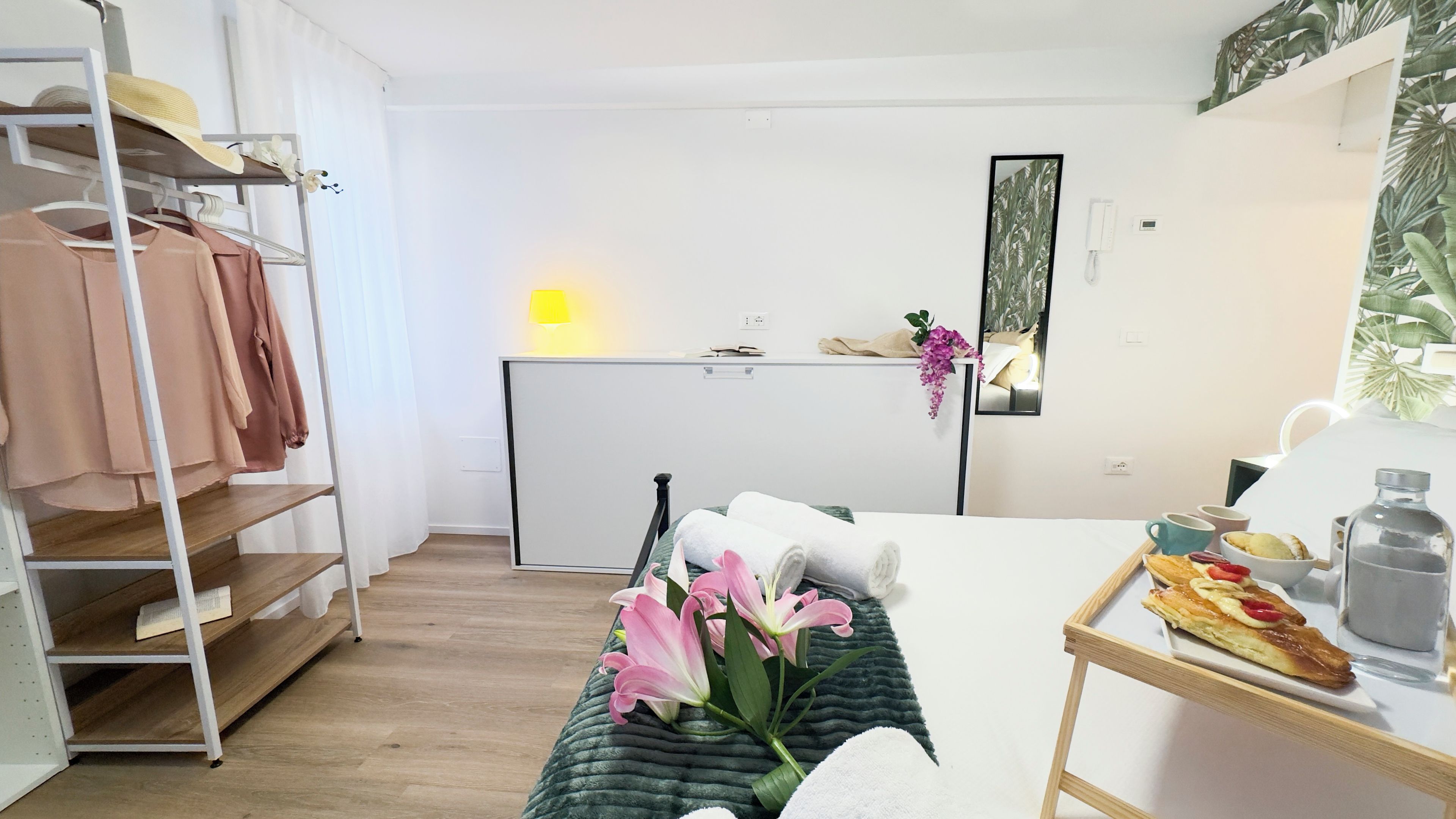 A modern, well-lit room with a bed, a bedside table with a tray of pastries, a mirror, and a wardrobe with hanging clothes.