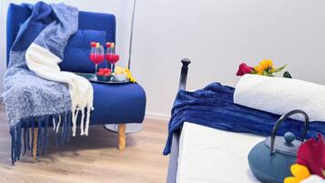 Une pièce avec un fauteuil bleu, une petite table sur laquelle se trouvent deux verres remplis d’une boisson rouge et des fruits, une serviette et une bouilloire.