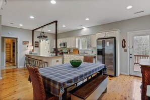 A modern kitchen with white cabinets, a stainless steel refrigerator, and a dining area with a plaid tablecloth and bench. - "TreeHouse" | Hot Tub | River Access | 20m to SNP (Front Royal)