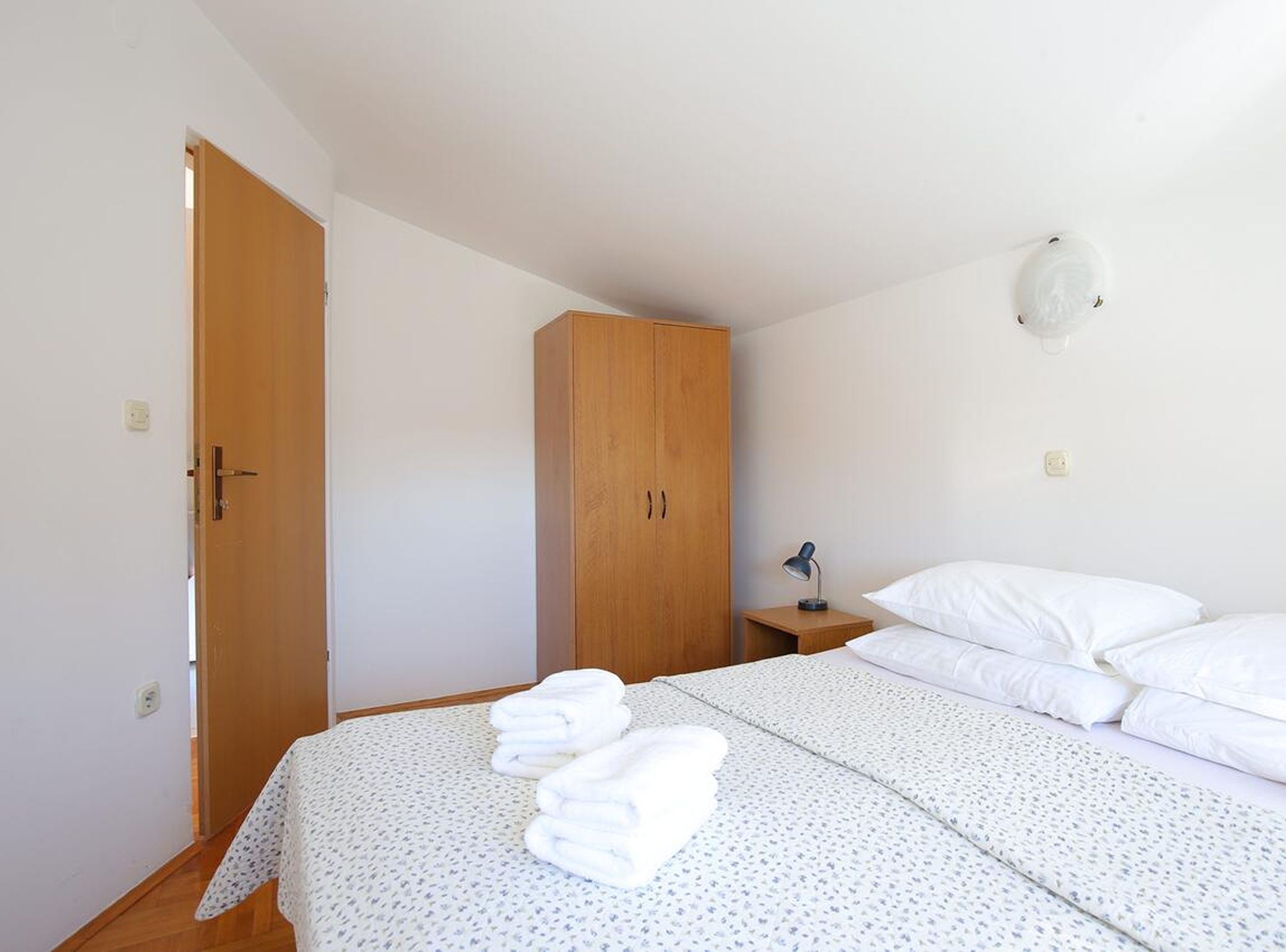 A bedroom with a bed, a wardrobe, a bedside table with a lamp, and a wall-mounted light fixture.