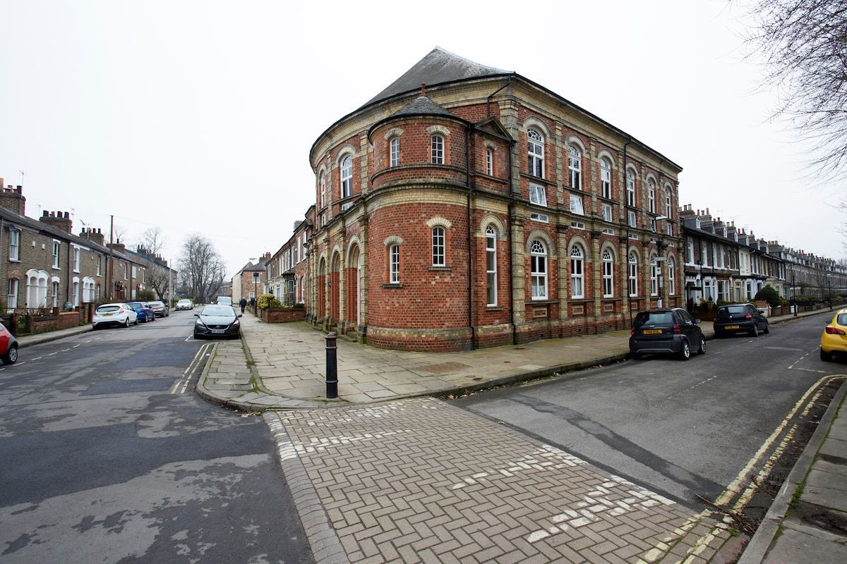 Un bâtiment en briques rouges, doté d’un coin arrondi, avec un trottoir pavé et des voitures garées dans une rue résidentielle.