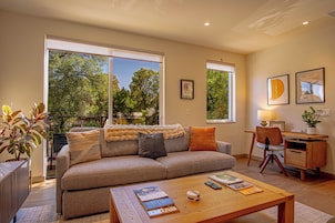 A living room with a sofa, coffee table, and a desk with a chair. There are windows with a view of trees and a balcony.