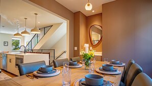 Zona pranzo con un tavolo lungo apparecchiato per quattro persone, una cucina sullo sfondo e una scala che conduce a un altro piano.