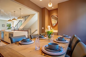 Espaço de refeições com uma mesa comprida posta para quatro pessoas, cozinha ao fundo e escada que leva a outro andar.