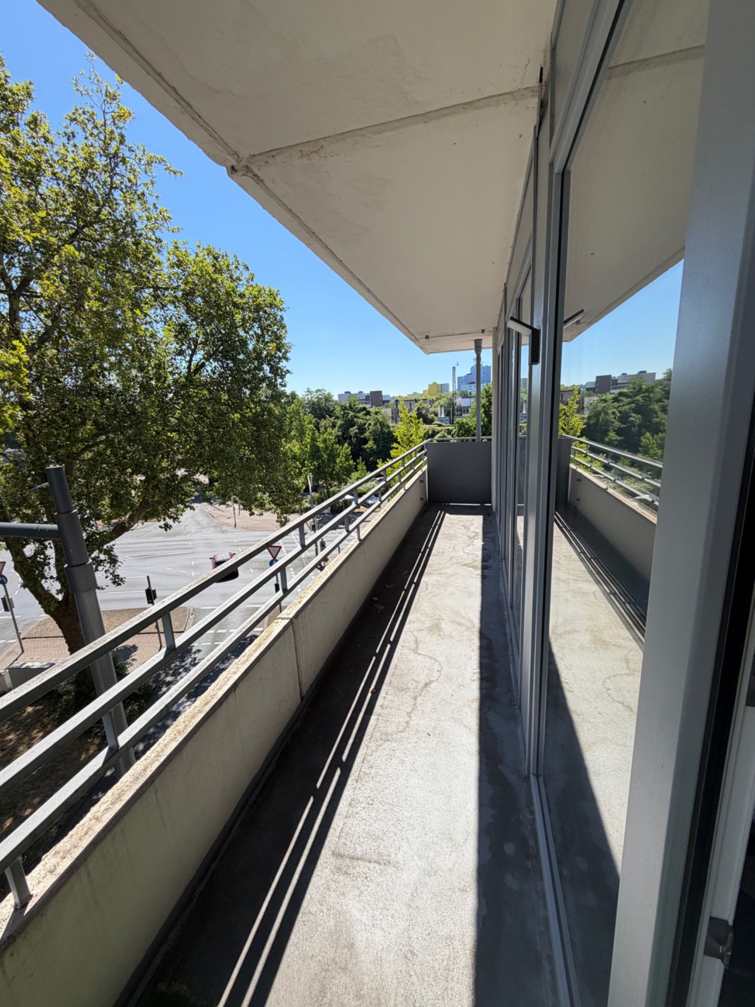 Ein Balkon mit Blick auf eine Straße und Bäume, mit Metallgeländern und einem Betonboden.