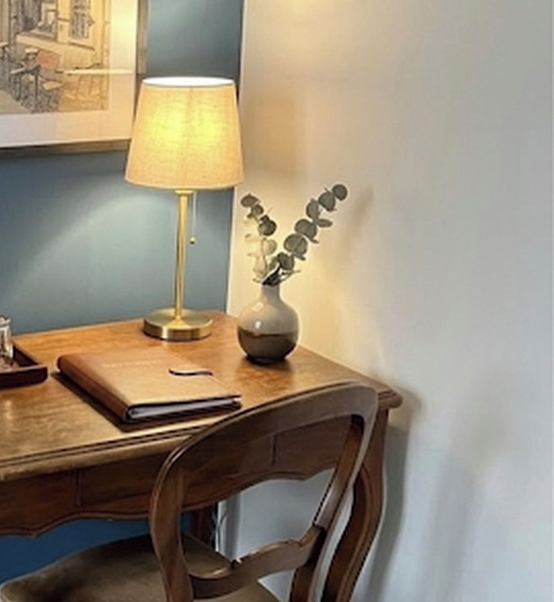 A wooden desk with a lamp, a vase with a plant, and a framed picture on the wall.