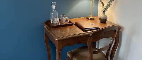A wooden desk with a chair, a framed picture on the wall, a lamp, and a tray with glasses and a bottle.