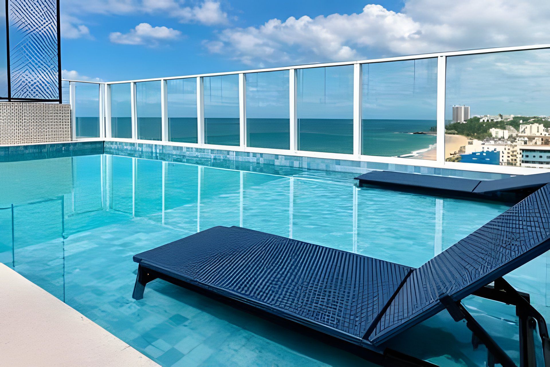 Piscina azul límpida com espreguiçadeiras, guarda-corpo de vidro e vista para o oceano e a paisagem urbana.