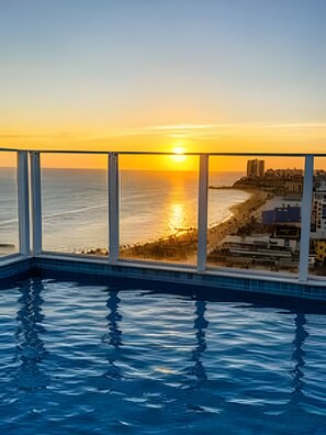 Uma piscina com vista para o pôr do sol sobre o oceano e um horizonte urbano ao fundo.