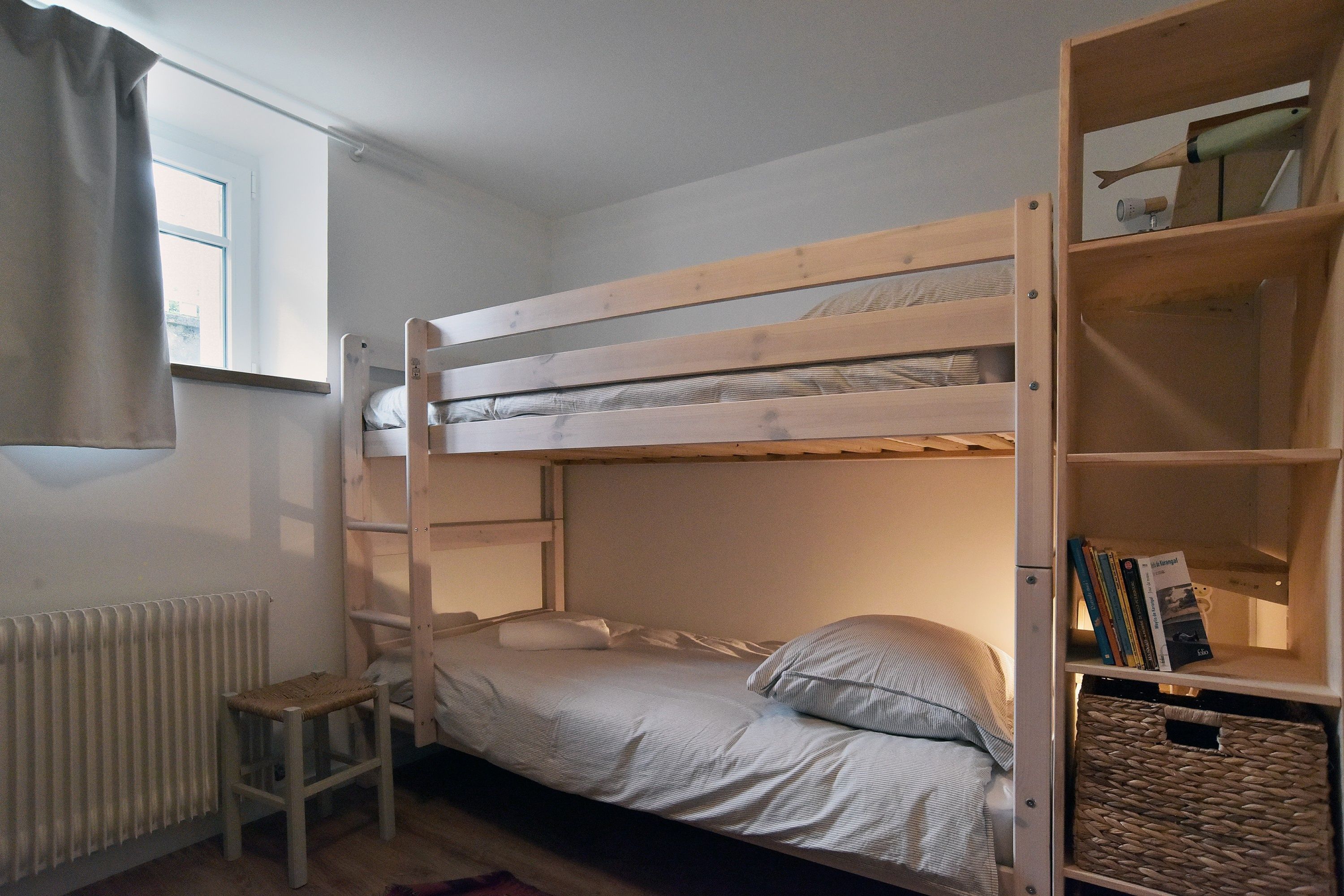 Una cama litera con escalera, una silla pequeña, un radiador y una estantería con libros y una cesta.